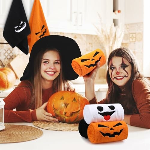 Two kids with Halloween face paint holding pumpkin-themed towels and a carved pumpkin in a kitchen.