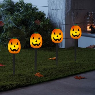Four glowing pumpkin lights on stakes in a garden.