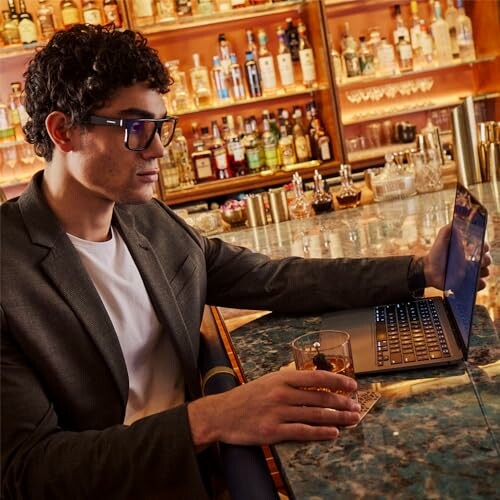 Man in glasses at bar with laptop and drink.